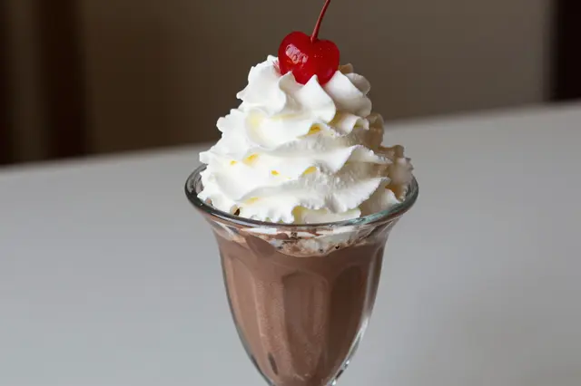 A glass chocolate sundae topped with swirled whipped cream and a whole red maraschino cherry.