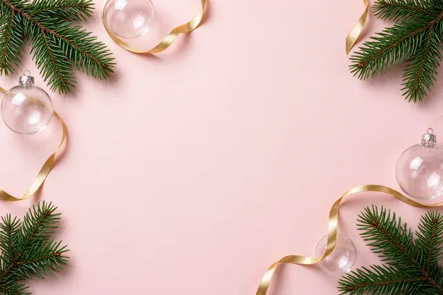 Overhead flat lay of a Christmas border with fir sprigs, clear baubles and gold ribbon on pale pink paper with empty center