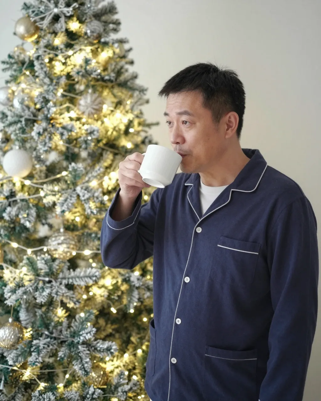 East Asian man in navy pajamas sips coffee next to a lit decorated Christmas tree