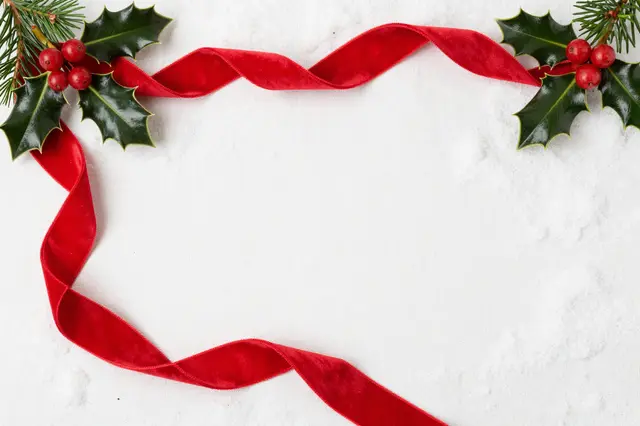 A curled red velvet ribbon and holly sprigs frame empty snowy white linen copy space