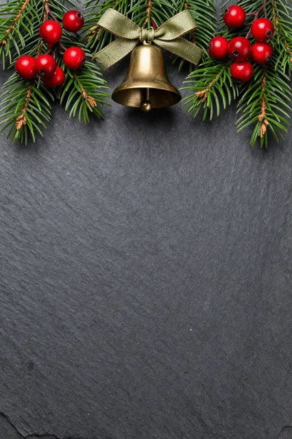 Festive cedar sprigs, red berries and a brass bell arranged at the top of a dark slate surface with empty copy space below