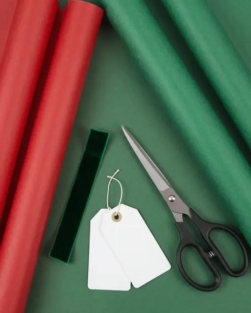Top-down view of red and green wrapping paper rolls, velvet ribbon, scissors and blank gift tags on a green surface