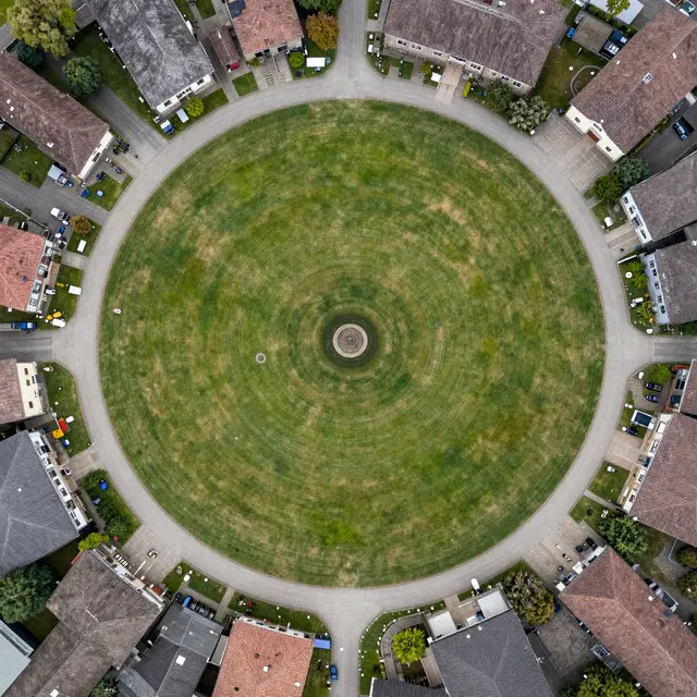 Top-down aerial view of a circular village green with radiating lanes and surrounding residential homes