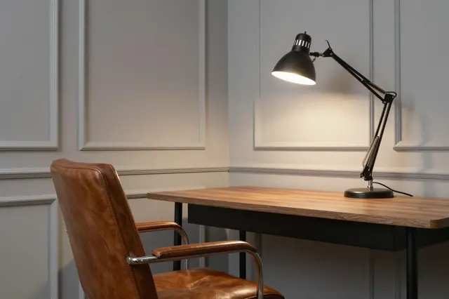 A lit desk lamp sits on a wooden work desk next to a brown leather office chair against paneled walls