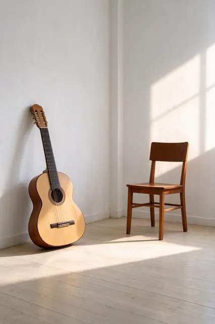 A nylon-string classical guitar leans in a white room corner beside a wooden chair in warm afternoon window sunlight