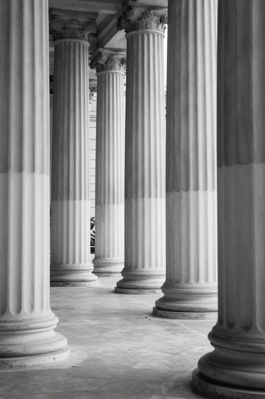 Black and white view of tall fluted classical columns in a sunlit empty stone portico