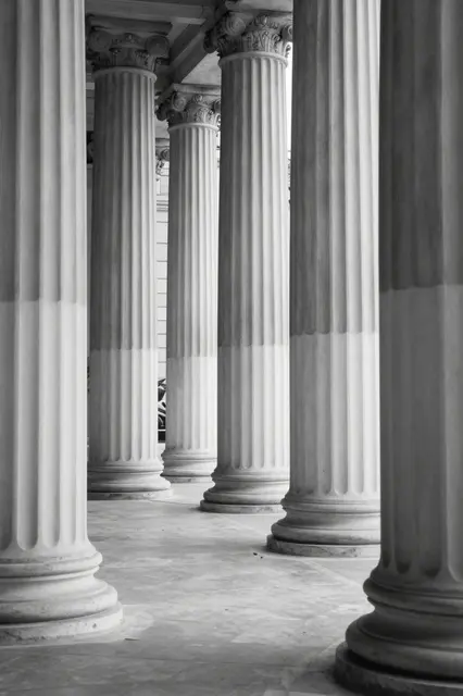 Black and white view of tall fluted classical columns in a sunlit empty stone portico
