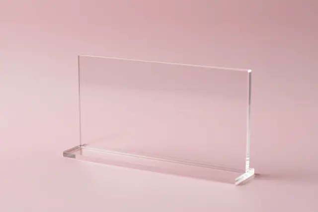 A clear rectangular acrylic display stand sits on a smooth pale pink backdrop with soft light refraction.