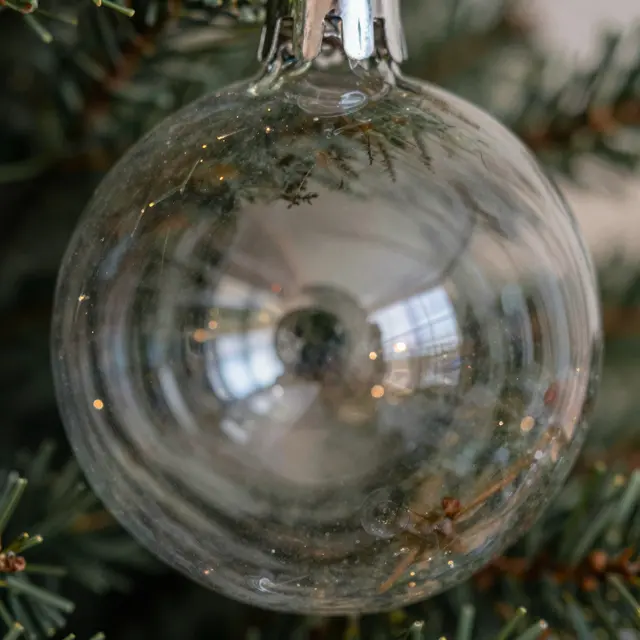 Close-up of a clear glass Christmas bauble hanging on a fir tree, reflecting warm festive bokeh lights.