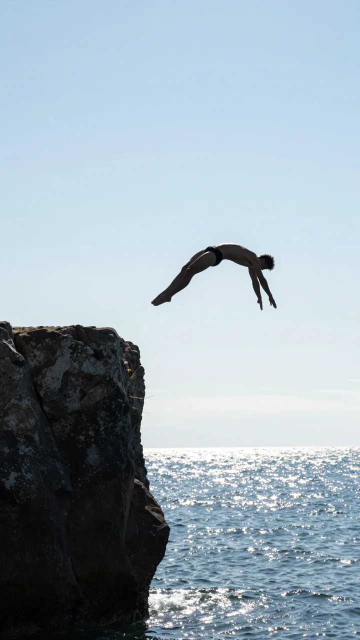 Silhouette of an adult cliff diver mid-air above sunlit ocean next to a rocky ledge