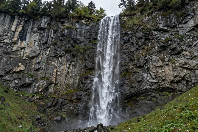 A tall cascading waterfall pours down a rugged sheer rock cliff into a grassy alpine valley