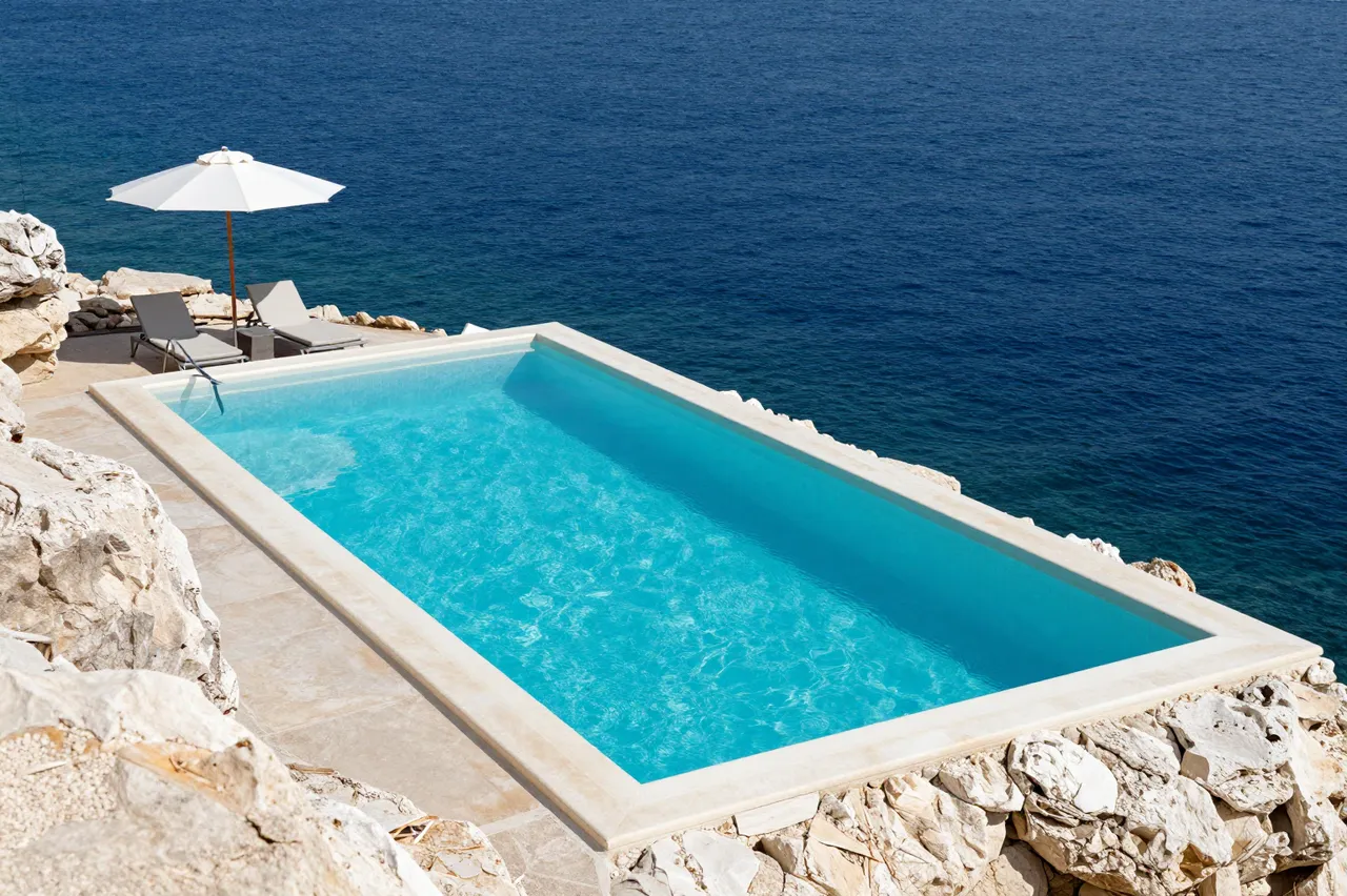 Empty rectangular infinity-style pool cut into pale rock overlooks a deep blue sea under midday sun, with a white parasol