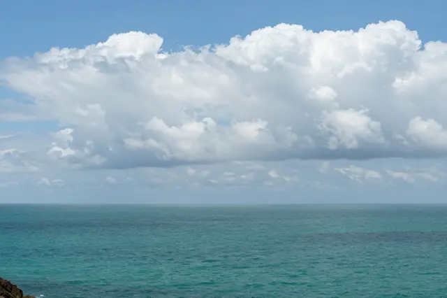 A wide view from a clifftop of turquoise sea below puffy white cumulus clouds in a bright blue sky.