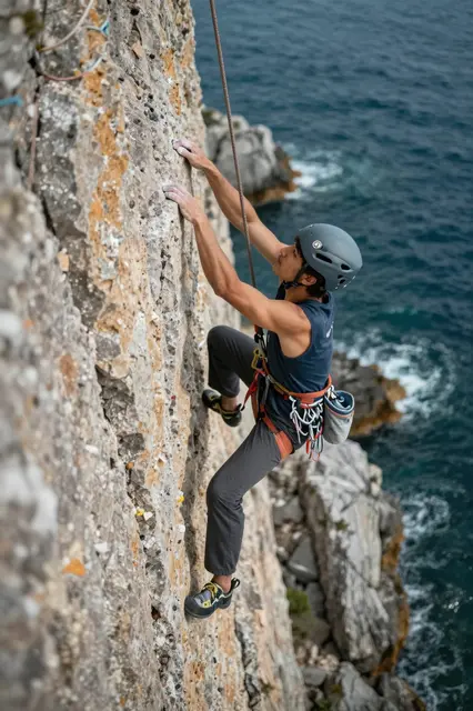 A male rock climber wearing a helmet ascends a steep ocean-side rock face above crashing sea waves.