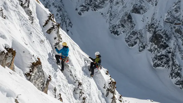 Two roped climbers in helmets ascend a steep snow-covered mountain ridge above a deep alpine valley