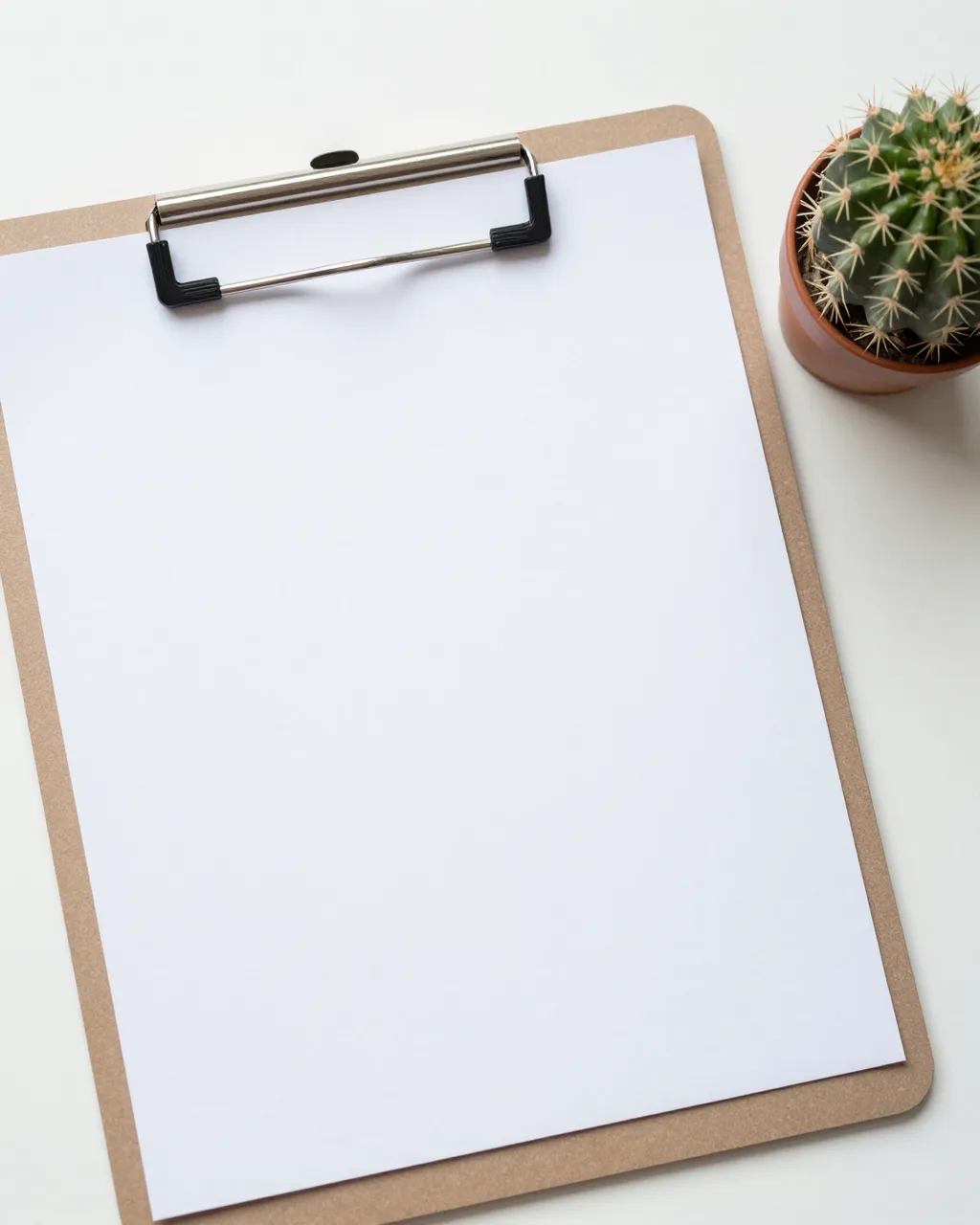 Top-down view of a blank paper clipboard next to a small potted cactus on a plain white desk surface.