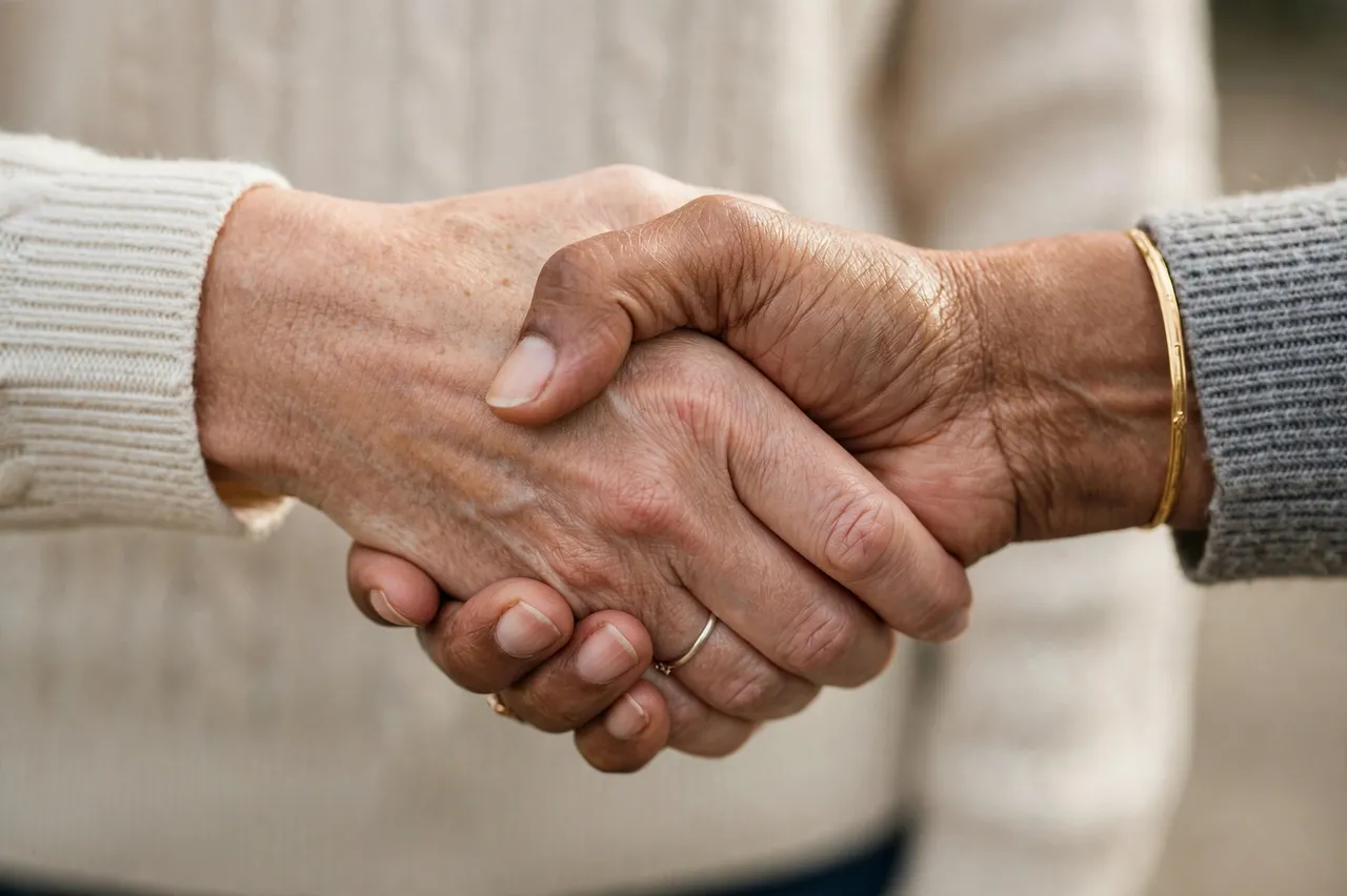 Close-up of a firm handshake between two older women wearing knit sweater sleeves, one light skinned and one brown skinned.