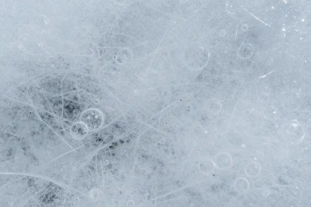 Frosted clear ice surface crisscrossed with fine cracks and dotted with trapped air bubbles