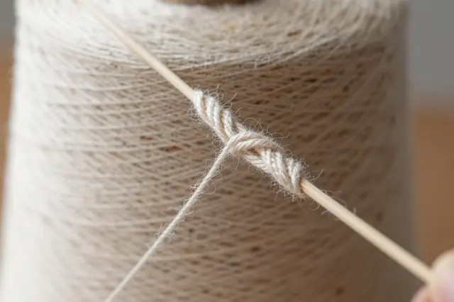 A close-up of a wooden spindle twisting natural wool thread in front of a yarn spool, with a small finger visible at the