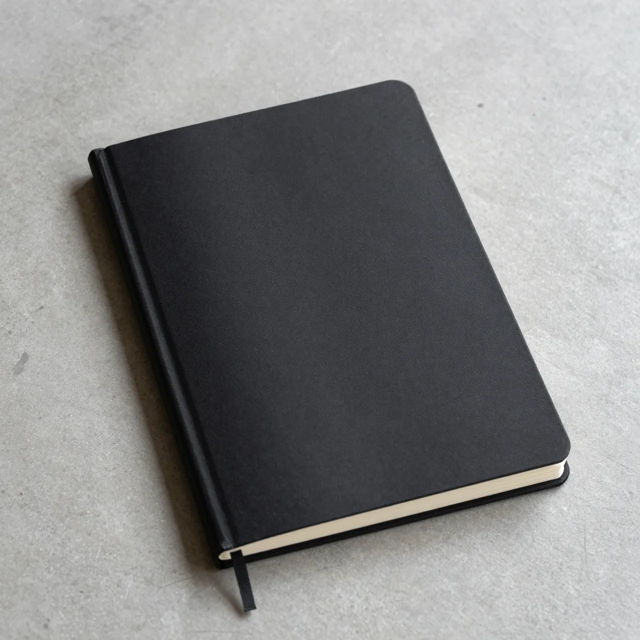 A plain closed black hardcover notebook rests flat on a smooth gray concrete surface