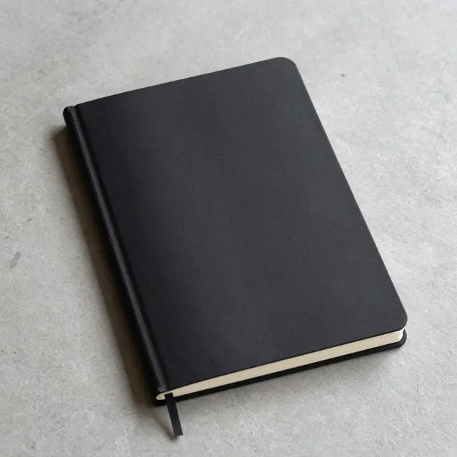 A plain closed black hardcover notebook rests flat on a smooth gray concrete surface