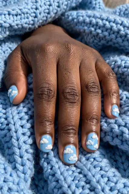 A Black person's hand with blue sky cloud nail art rests on a light blue knit blanket