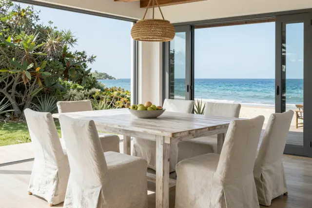 Bright coastal dining room with whitewashed table, slipcovered chairs, and ocean view through open sliding doors