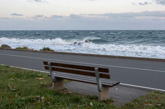 A wooden roadside bench faces choppy stormy ocean waves under a dusky overcast sky