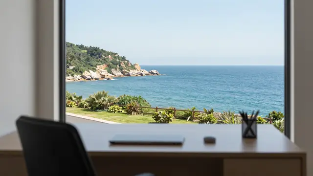 Light wood desk with office supplies sits before a large window overlooking a bright turquoise coastal ocean view