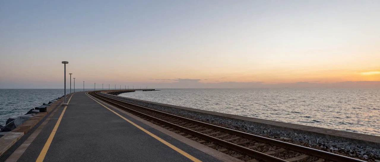 A long curving railway platform beside the calm sea at soft gradient sunset, viewed from the near end of the structure.