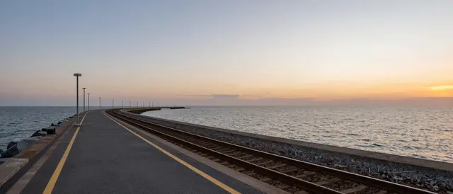 A long curving railway platform beside the calm sea at soft gradient sunset, viewed from the near end of the structure.