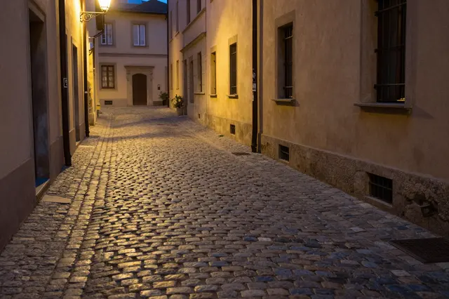 A quiet cobblestone European alley is lit by warm glowing street lamps after dark