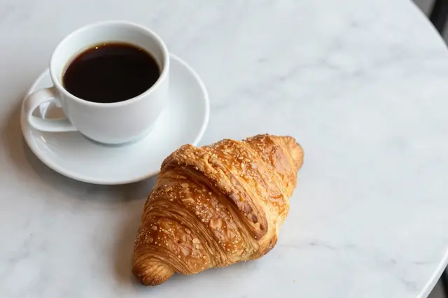 Top-down view of a black coffee cup on a saucer next to a sesame-topped croissant on a light marble table with empty copy