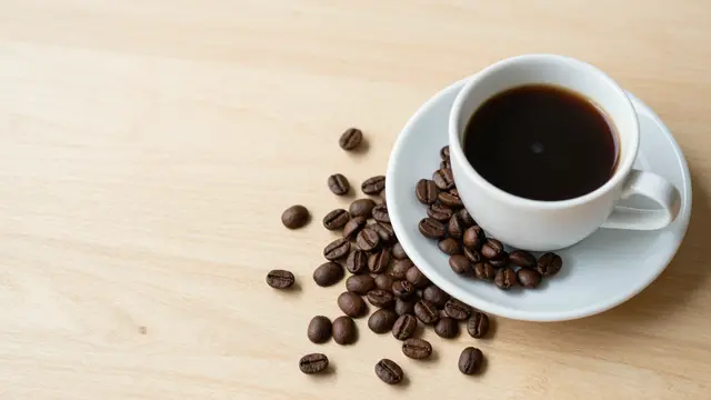 Top view of a white coffee cup with black coffee, scattered roasted beans, and wide empty light wooden table copy space