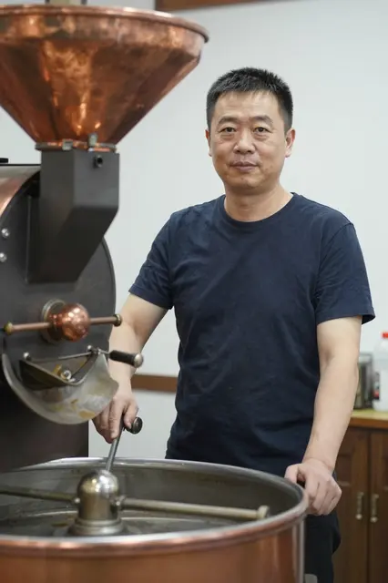 East Asian man in his forties stands beside a copper coffee roasting machine in a small roastery