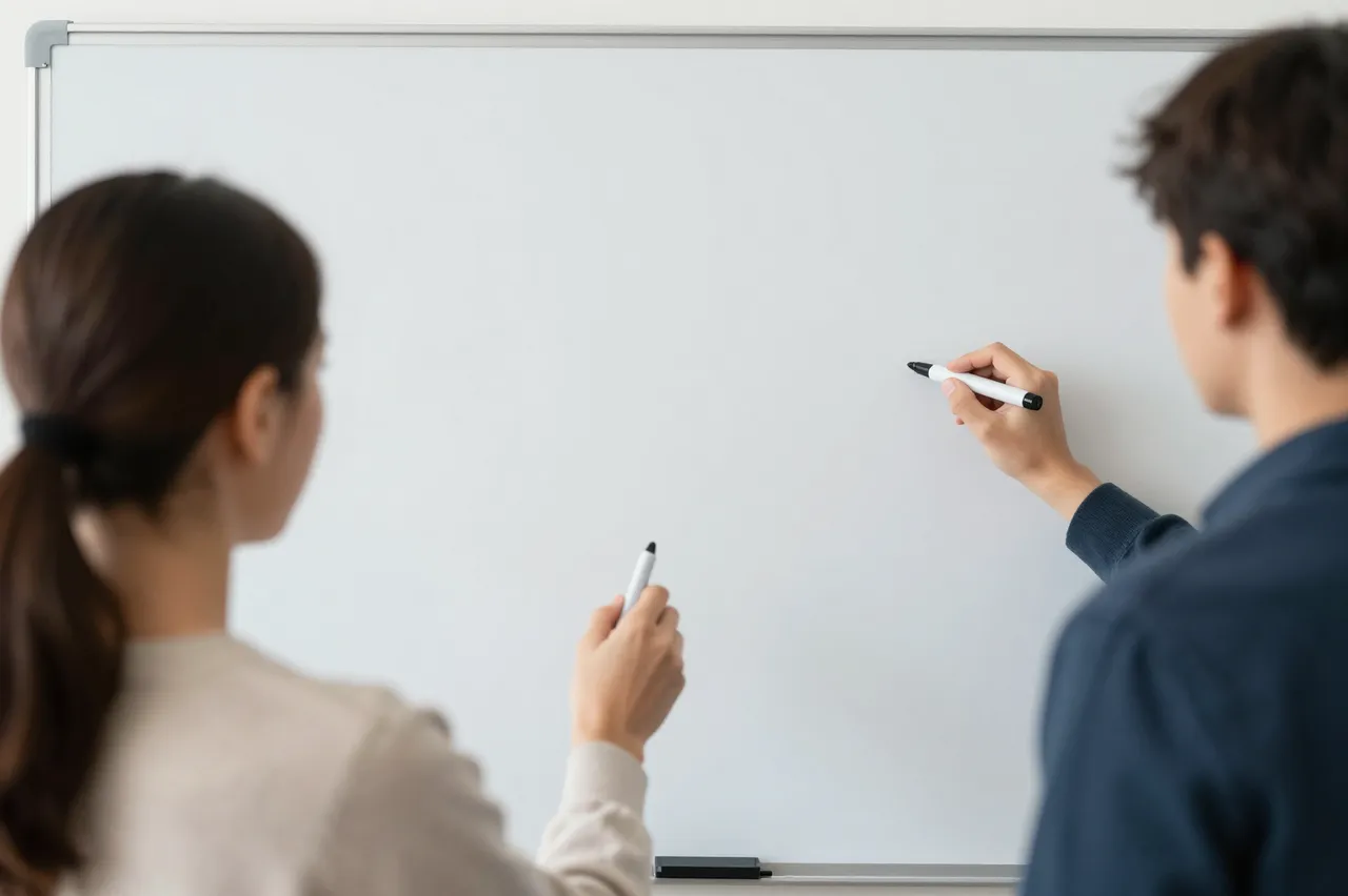 Two people viewed from behind hold dry erase markers while standing in front of a blank office whiteboard.