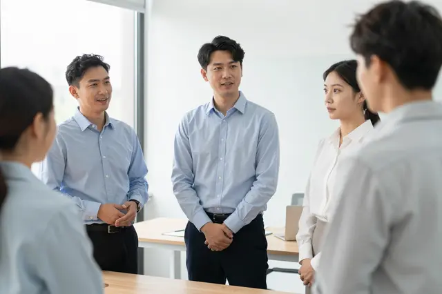 A group of adult office coworkers gather for a casual standing meeting in a bright modern office space.
