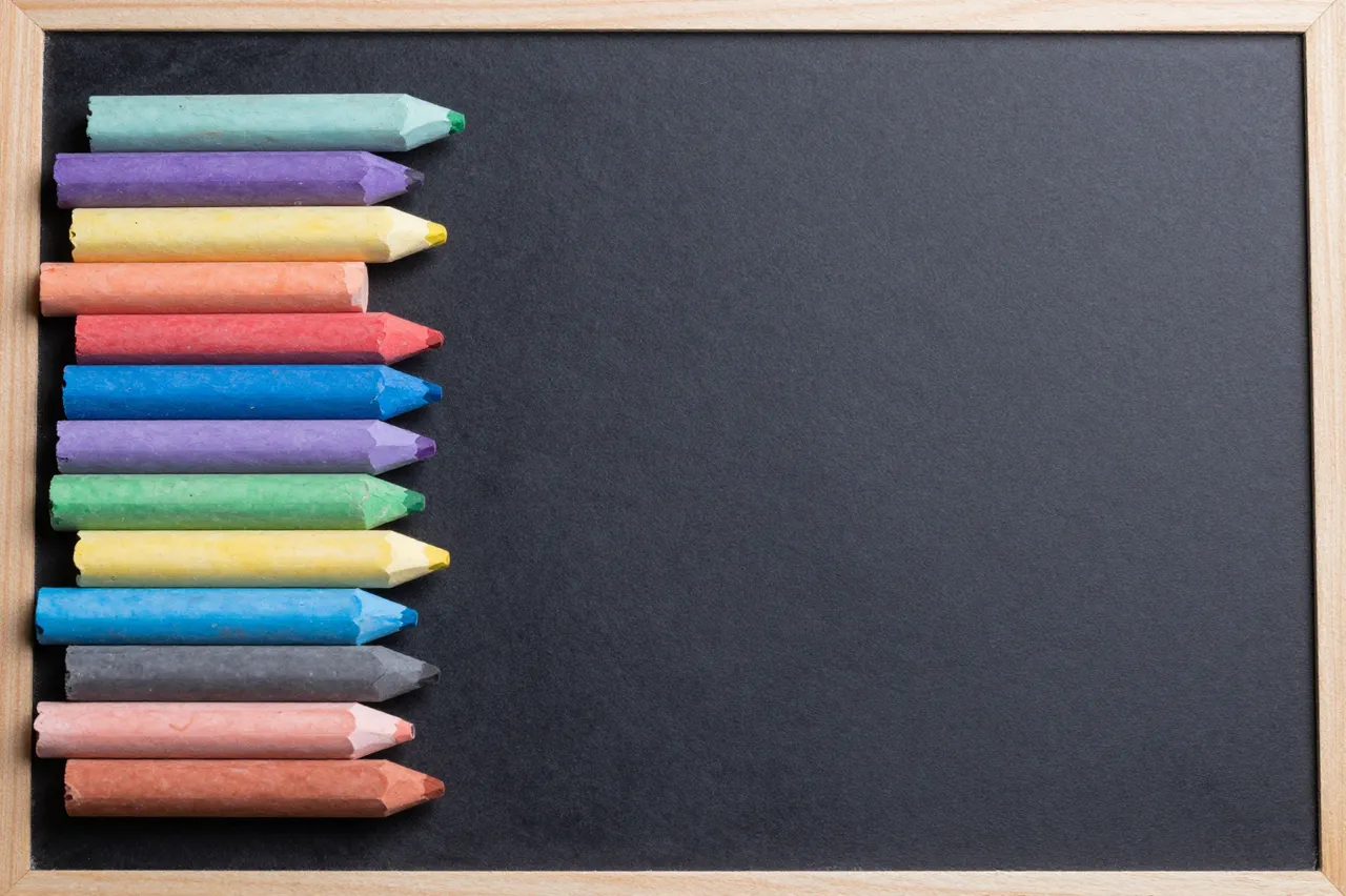 Top view of assorted colored chalk sticks arranged along the left edge of a blank wooden-framed chalkboard