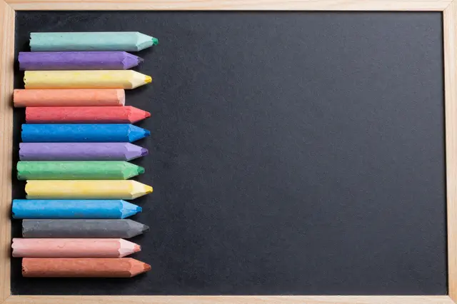 Top view of assorted colored chalk sticks arranged along the left edge of a blank wooden-framed chalkboard