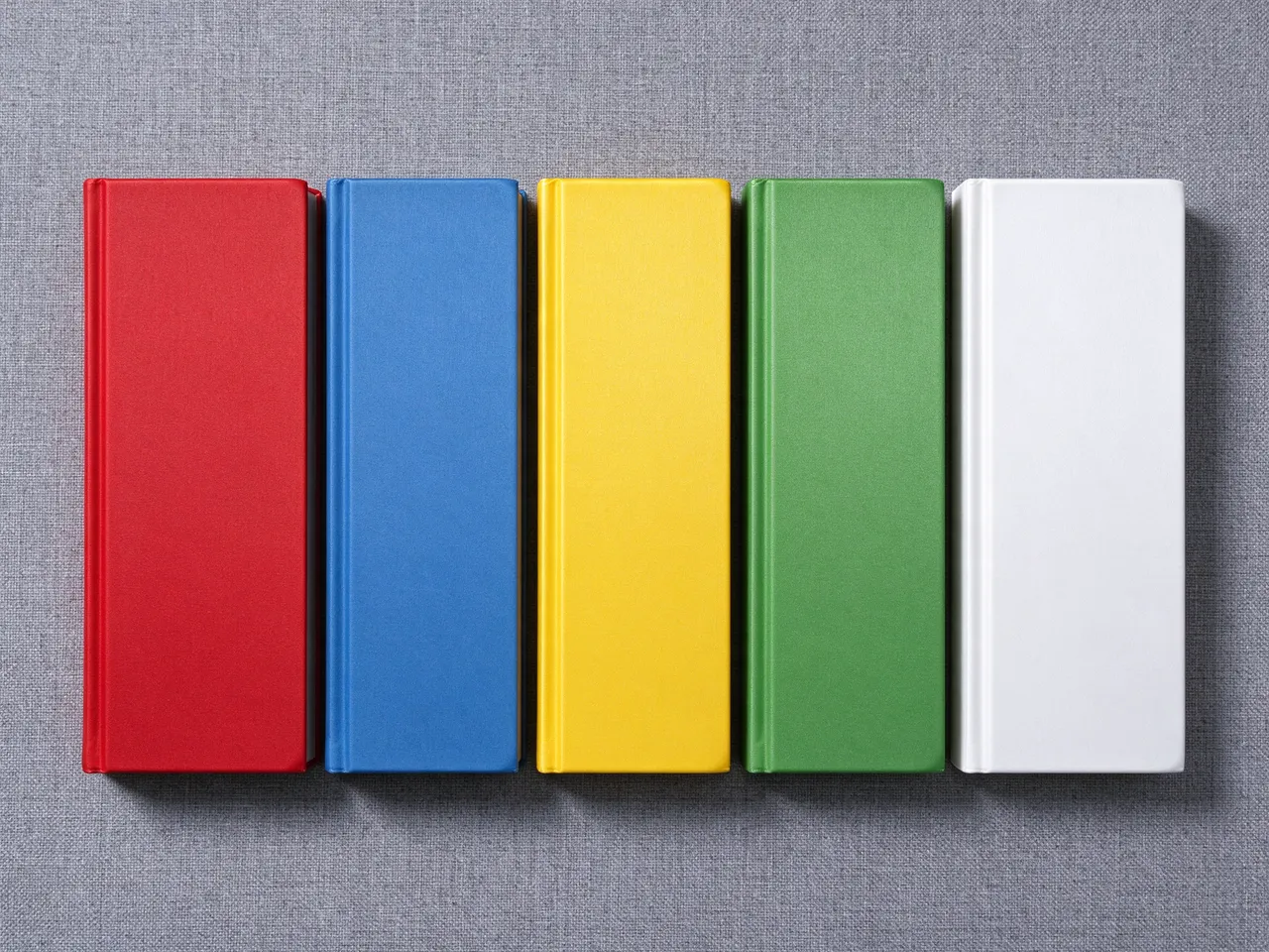 Five plain red, blue, yellow, green, and white closed hardcover books rest in a row on gray linen fabric, viewed from above.