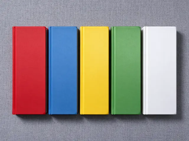 Five plain red, blue, yellow, green, and white closed hardcover books rest in a row on gray linen fabric, viewed from above.