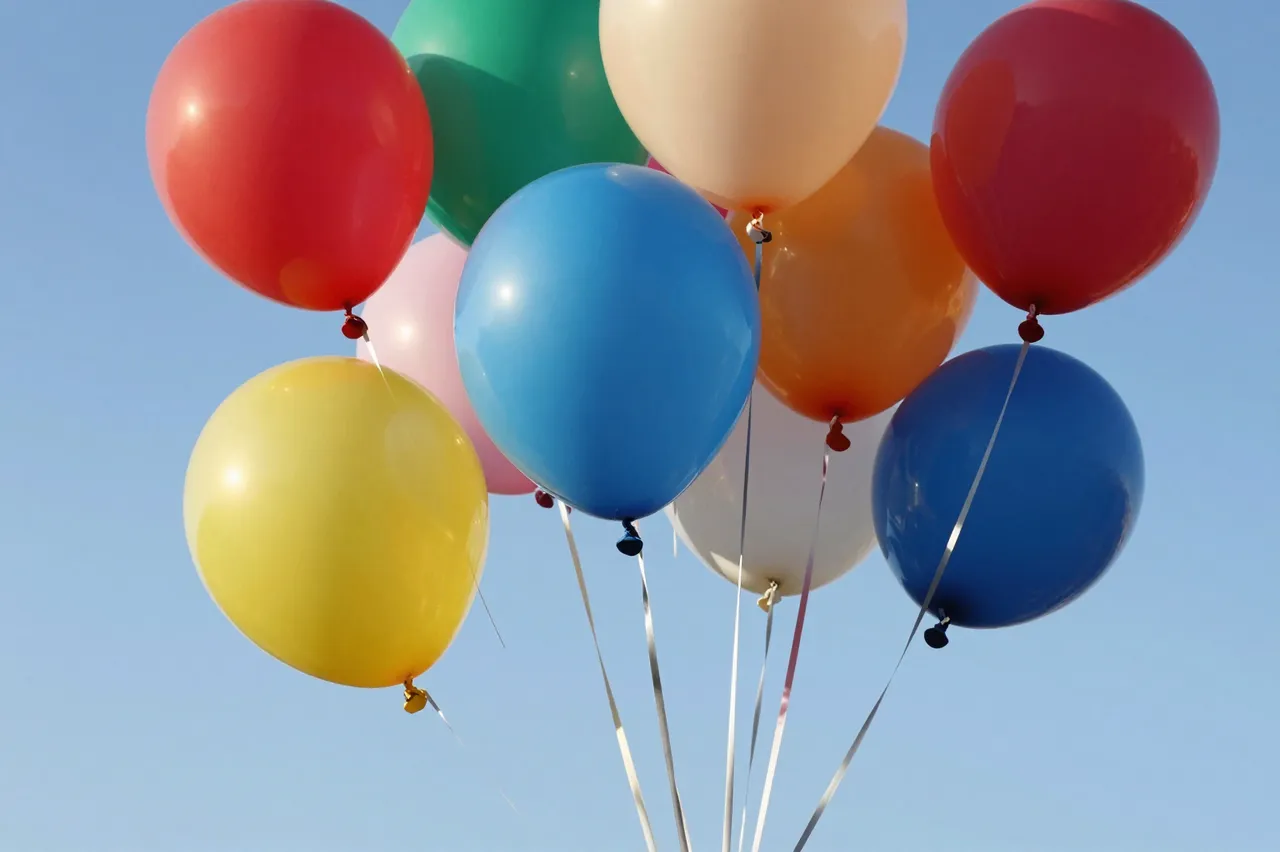 A cluster of bright multicolored party balloons floats against a clear light blue sky.