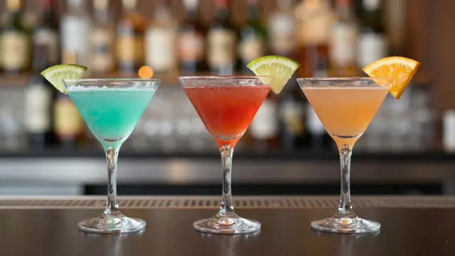 Three martini glasses of blue, red and orange cocktails sit in a row on a dark bar top with warm blurred bokeh behind.