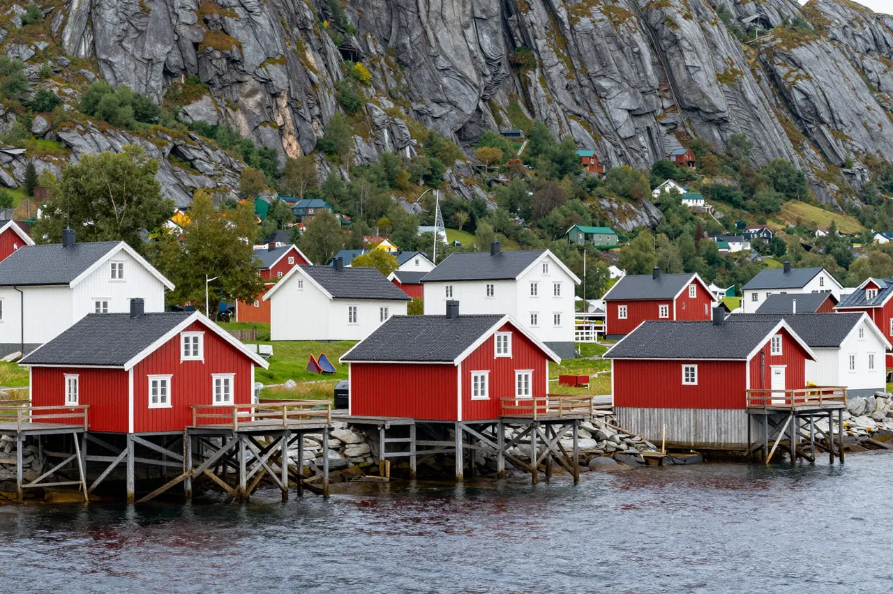 Red and white stilted fishing houses line a rocky nordic fjord beneath a steep mountain backdrop