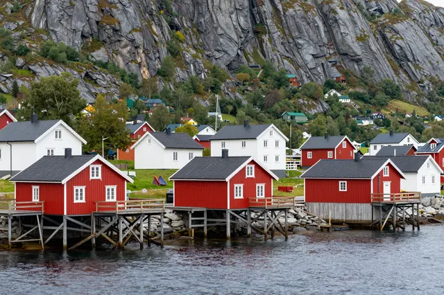 Red and white stilted fishing houses line a rocky nordic fjord beneath a steep mountain backdrop