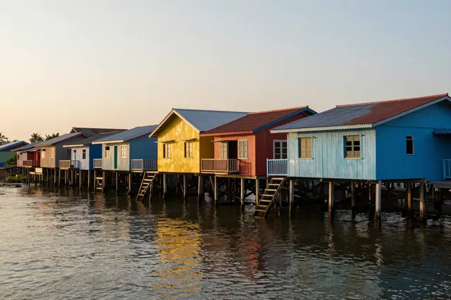 A row of brightly painted wooden stilt houses lines a calm river under a soft sunset sky