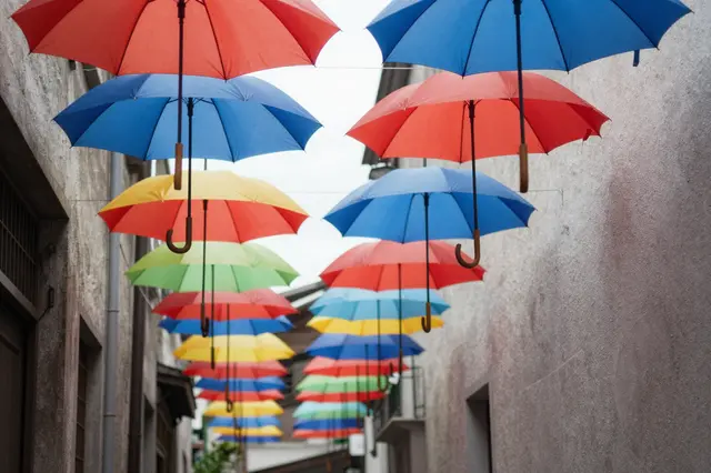 Rows of bright colorful umbrellas hang suspended above a narrow plastered alleyway between old buildings.