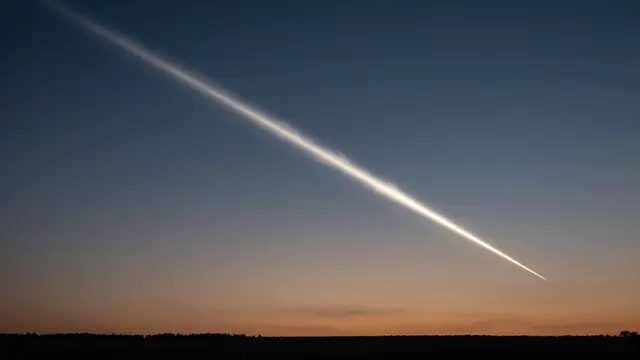 A bright glowing comet trail cuts diagonally across a gradient dusk sky above a dark silhouetted horizon