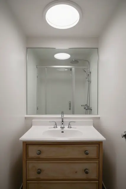 Small bathroom with wooden vanity, large mirror, round ceiling light and shower reflected in glass
