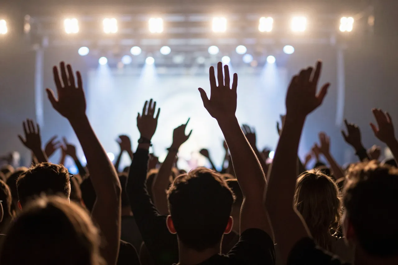 Rear view of a concert audience with raised hands lit by bright overhead stage lights